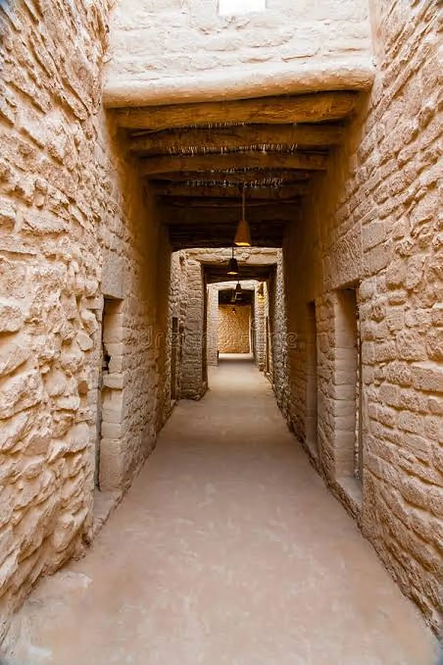 A shaded lane between stone-and-mudbrick walls in AlUla Old Town, roofed with wooden beams and hanging lamps
