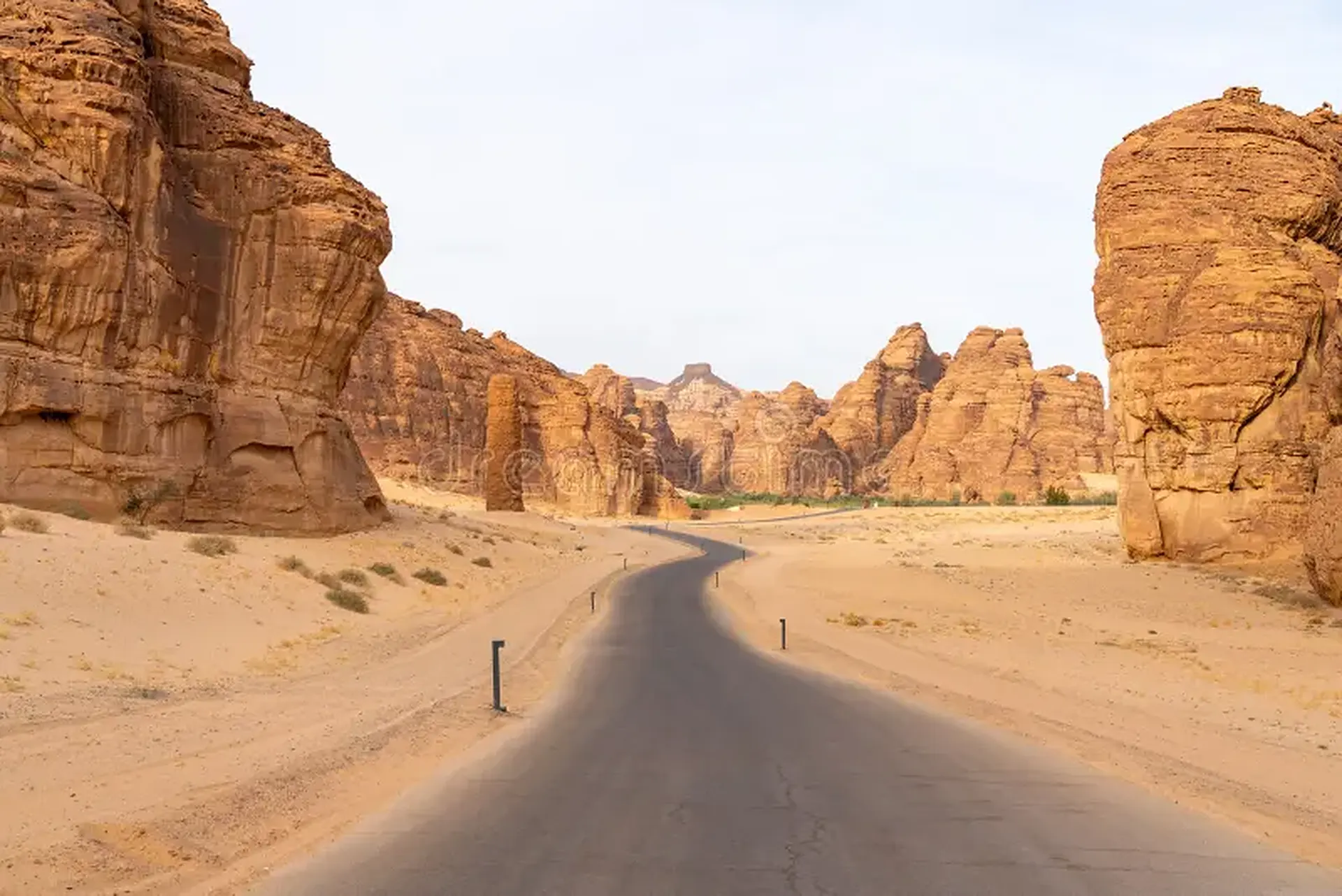 A paved road winding between towering sandstone rock formations and pale desert sand near AlUla