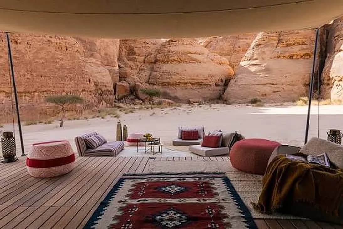 An open-sided desert camp lounge with rugs, cushions and low seating facing sandstone cliffs