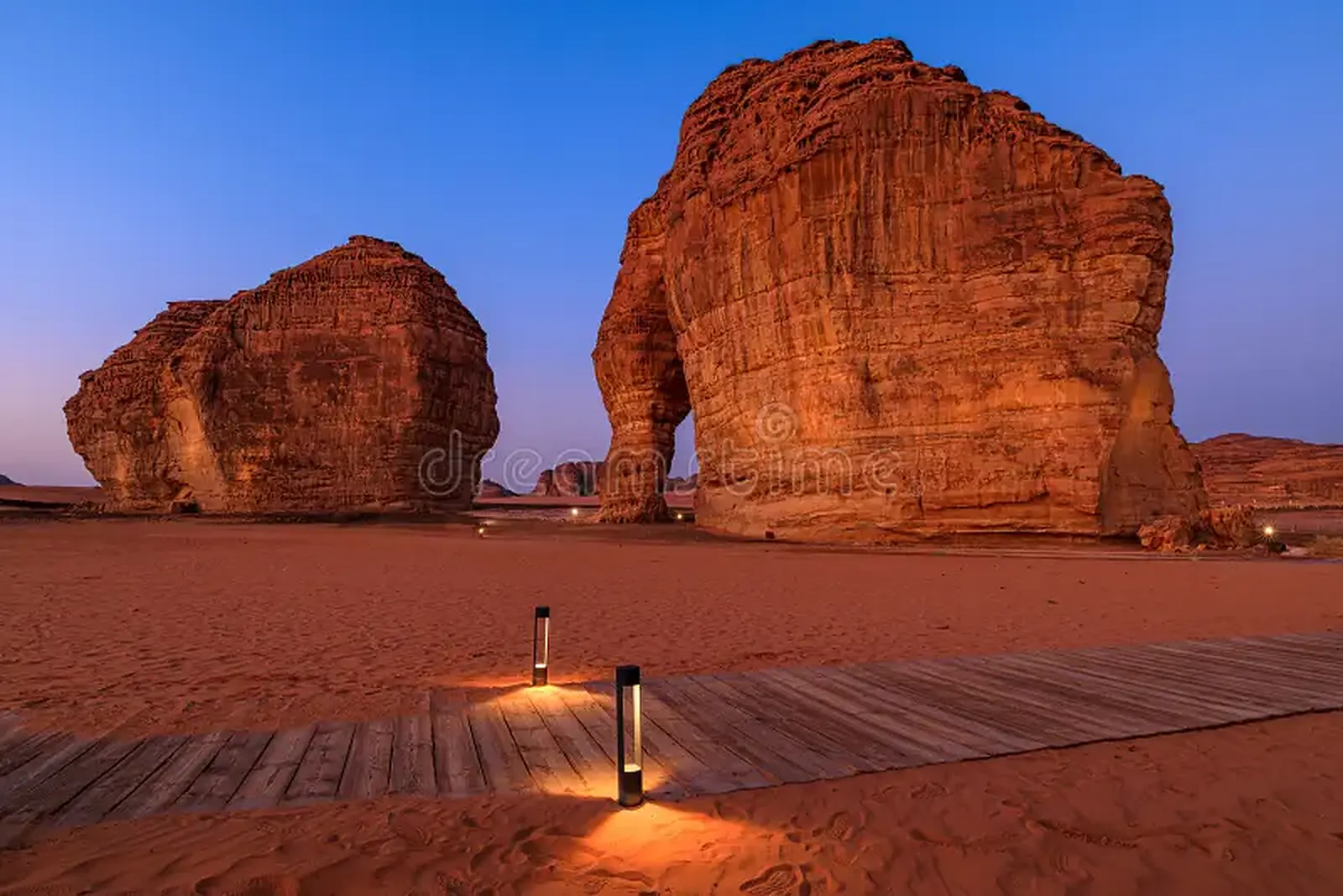 Elephant Rock (Jabal AlFil), a sandstone formation with a natural arch, lit at dusk above a wooden walkway with low lights