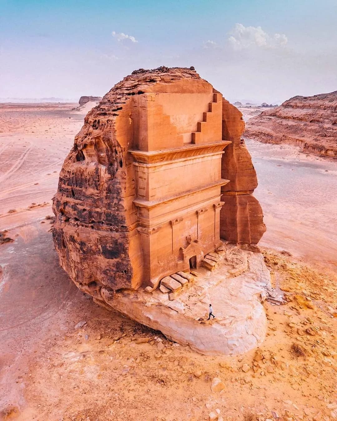 Qasr al-Farid, a monumental tomb carved into a freestanding sandstone outcrop at Hegra, with a visitor standing on the rock below for scale